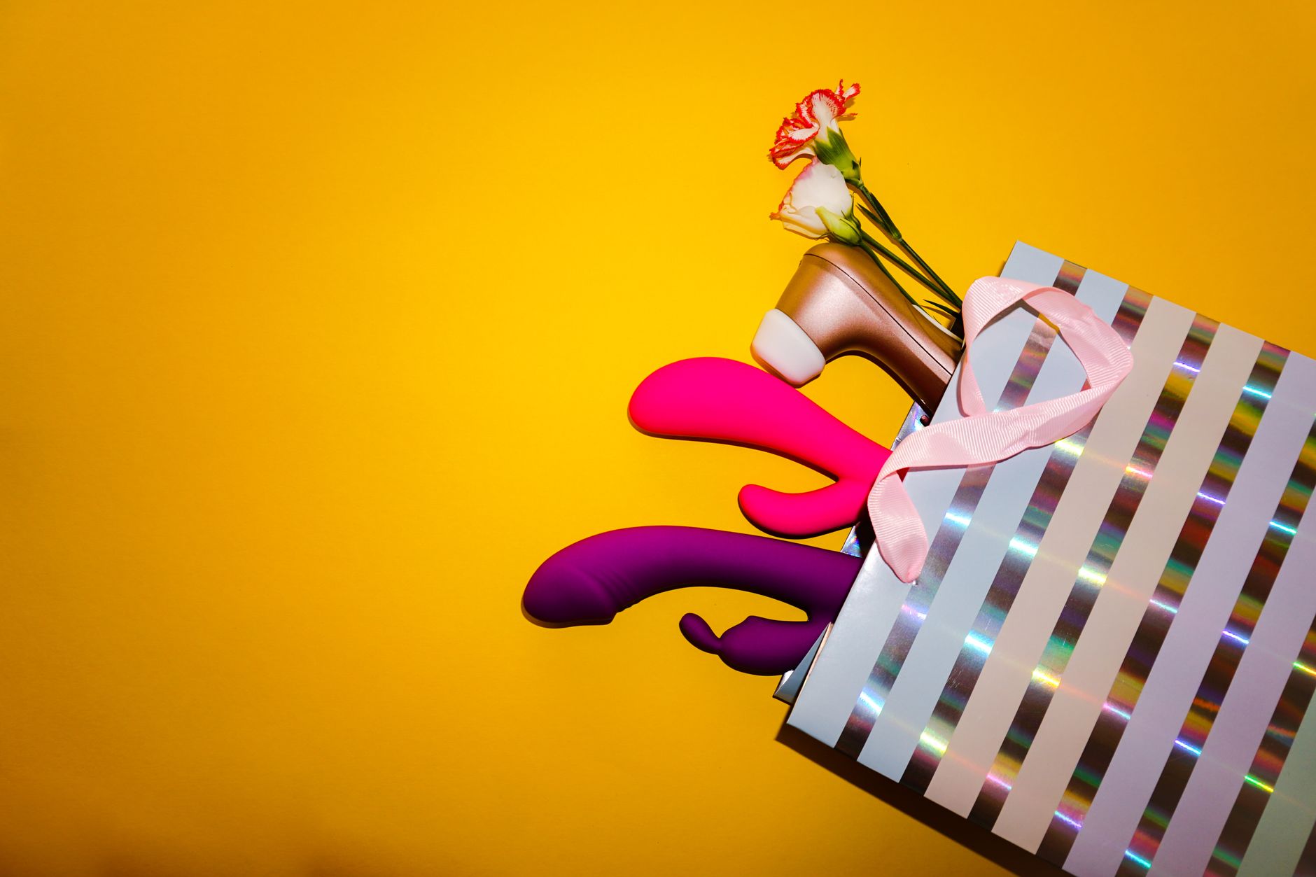Colorful clitoral vibrators with fresh flowers in a holographic gift bag against a bright yellow background.